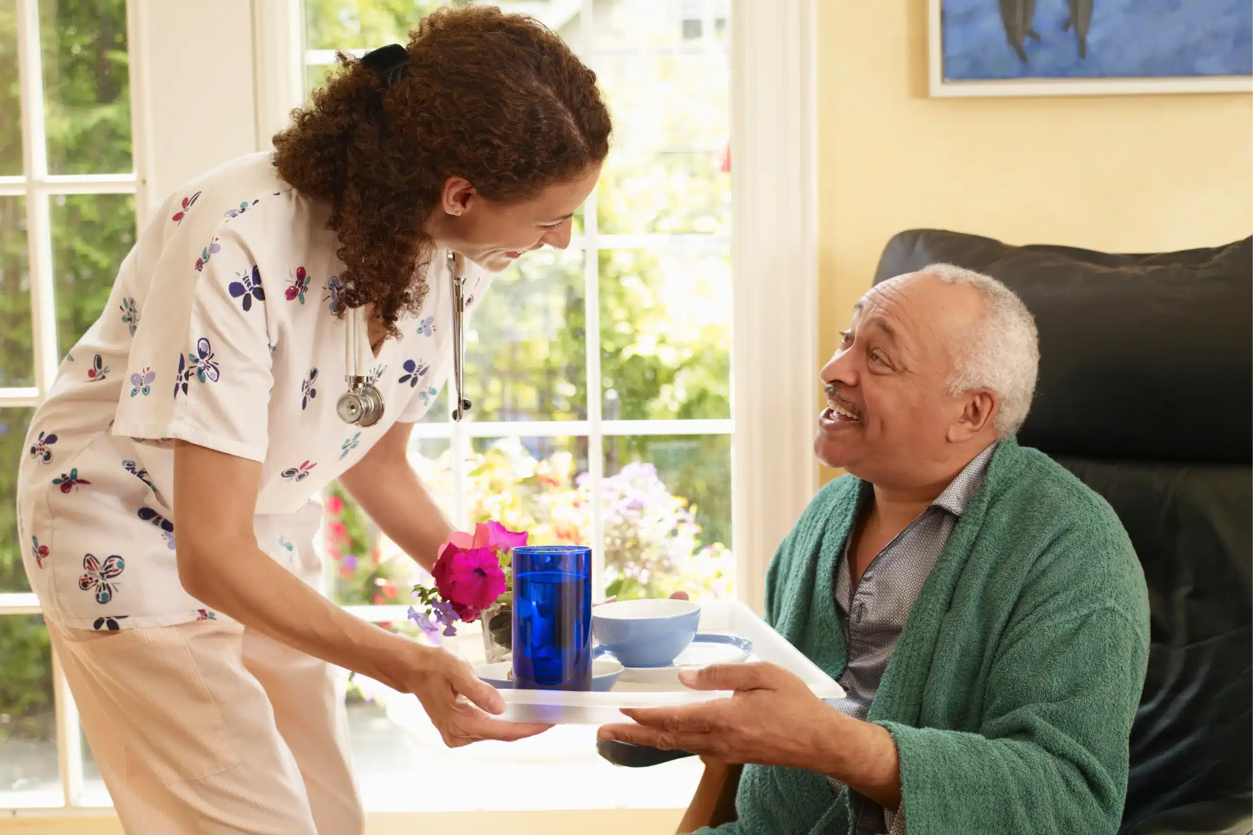 A woman serves a cup of tea on a tray to an elderly man, both smiling in a cozy setting.