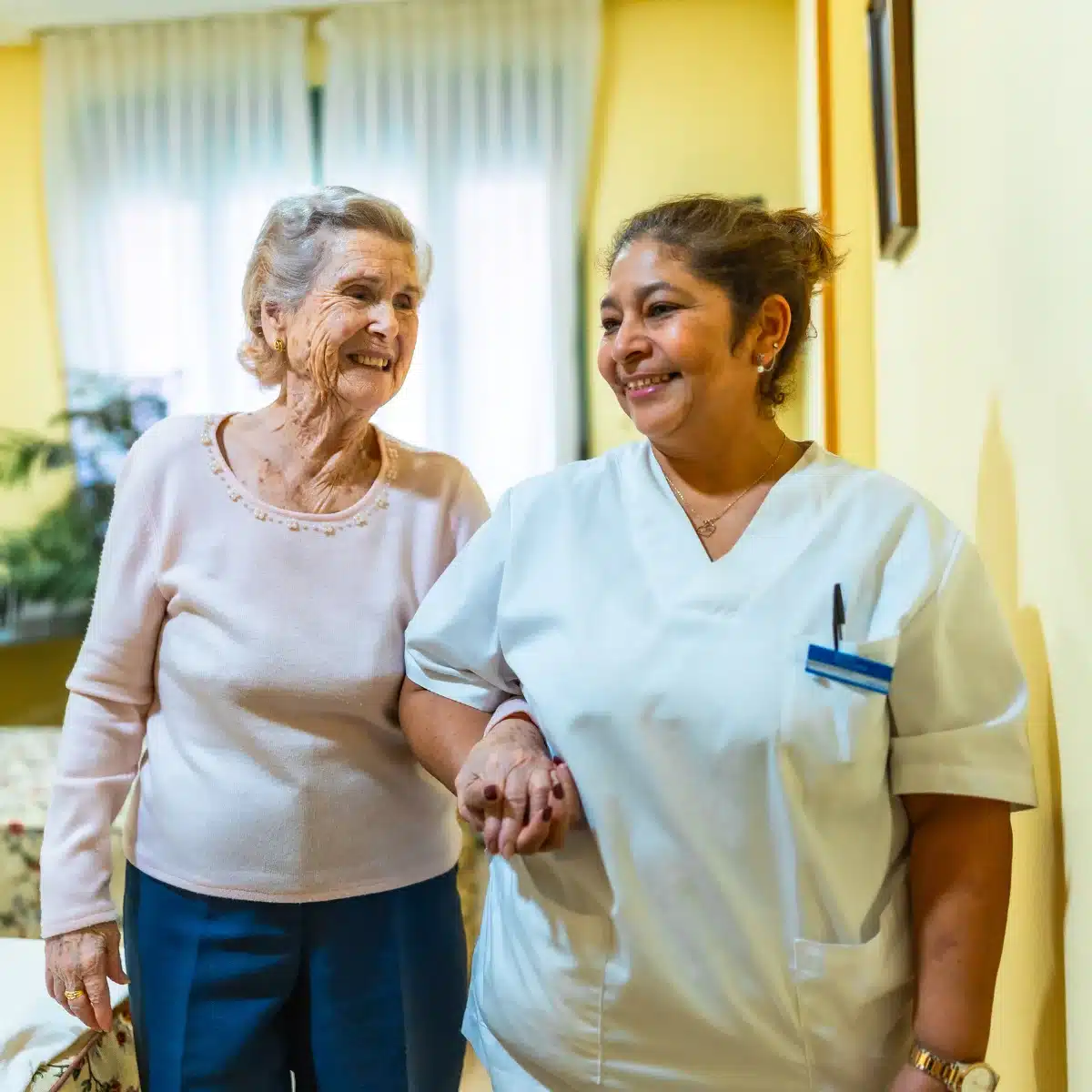 A nurse and an elderly woman stand together in a brightly lit hallway, engaged in conversation.